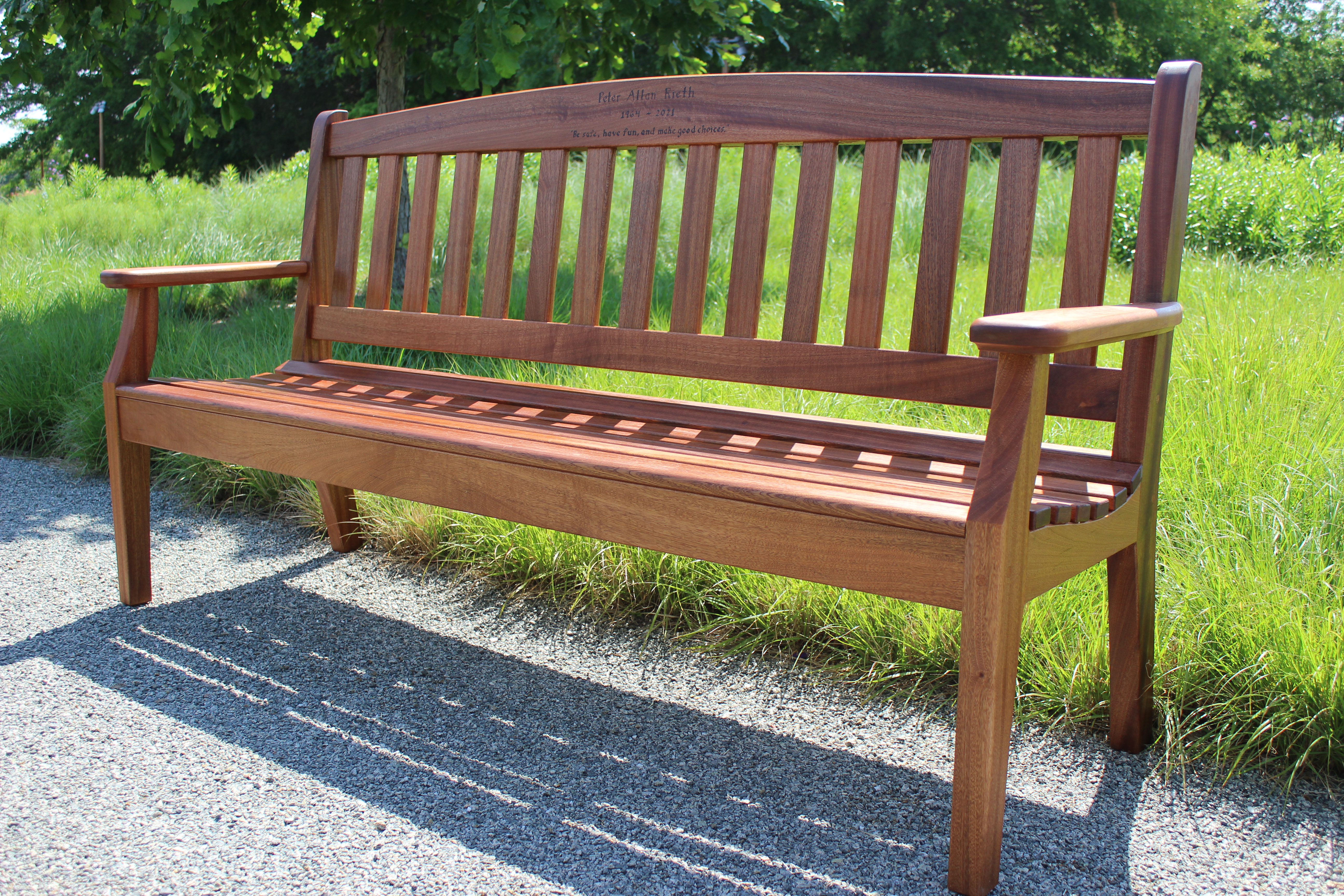A wooden bench with a slatted backrest and seat, placed on a gravel path surrounded by tall grass.