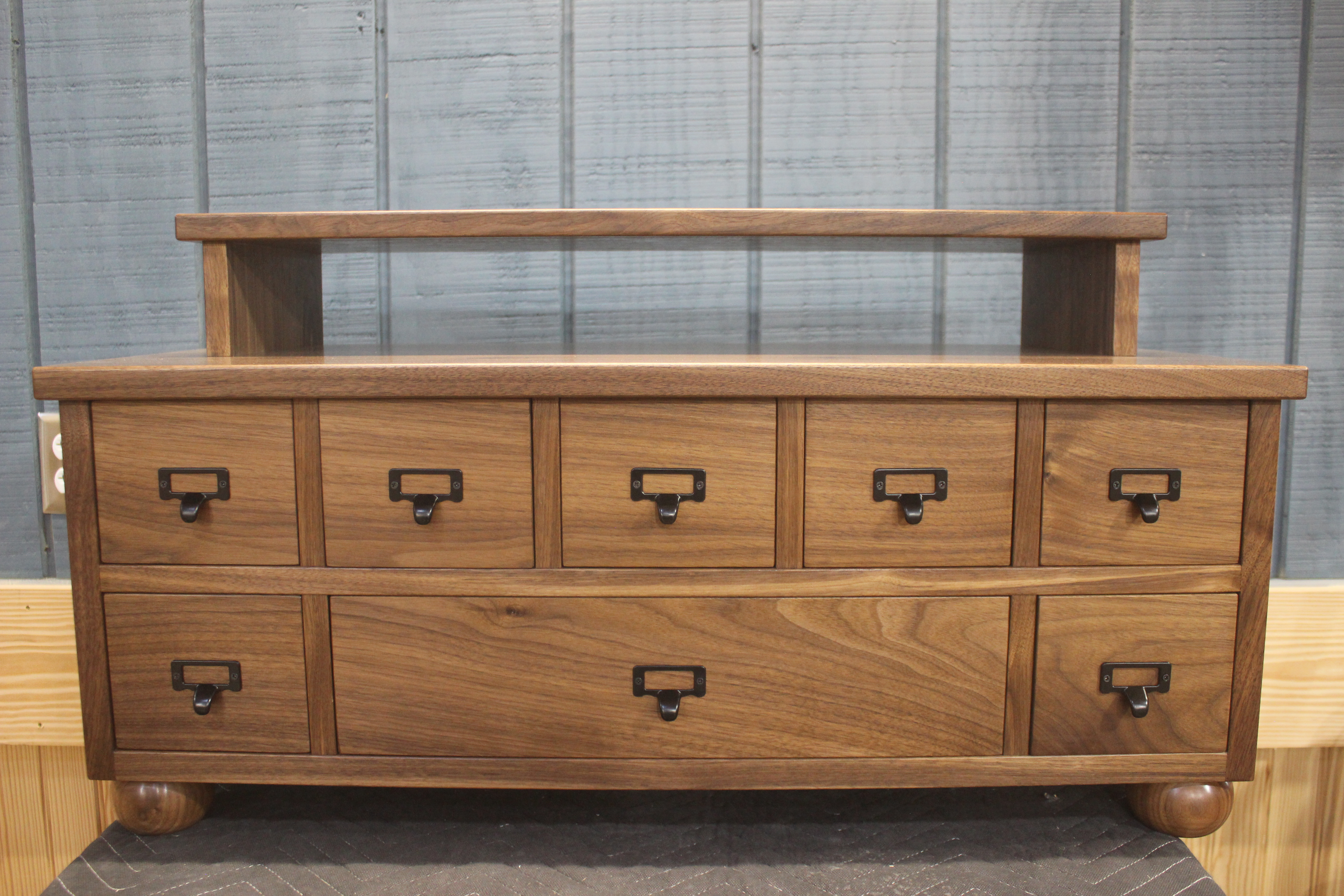 A wooden dresser with multiple drawers, featuring a smooth top and black metal handles, set against a gray wall.