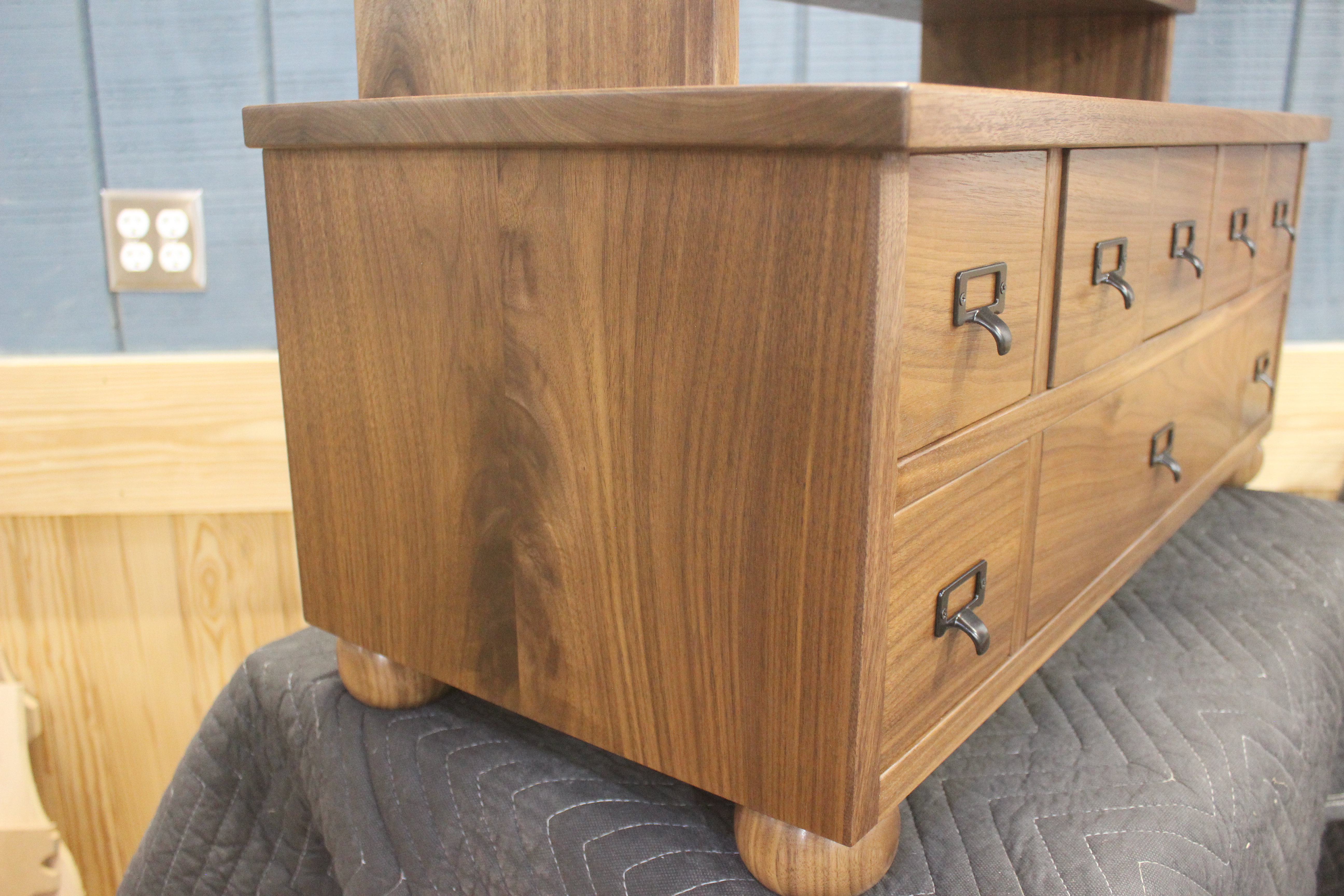 A close-up view of a wooden chest of drawers, showcasing its rich walnut finish, rounded feet, and black metal handles.