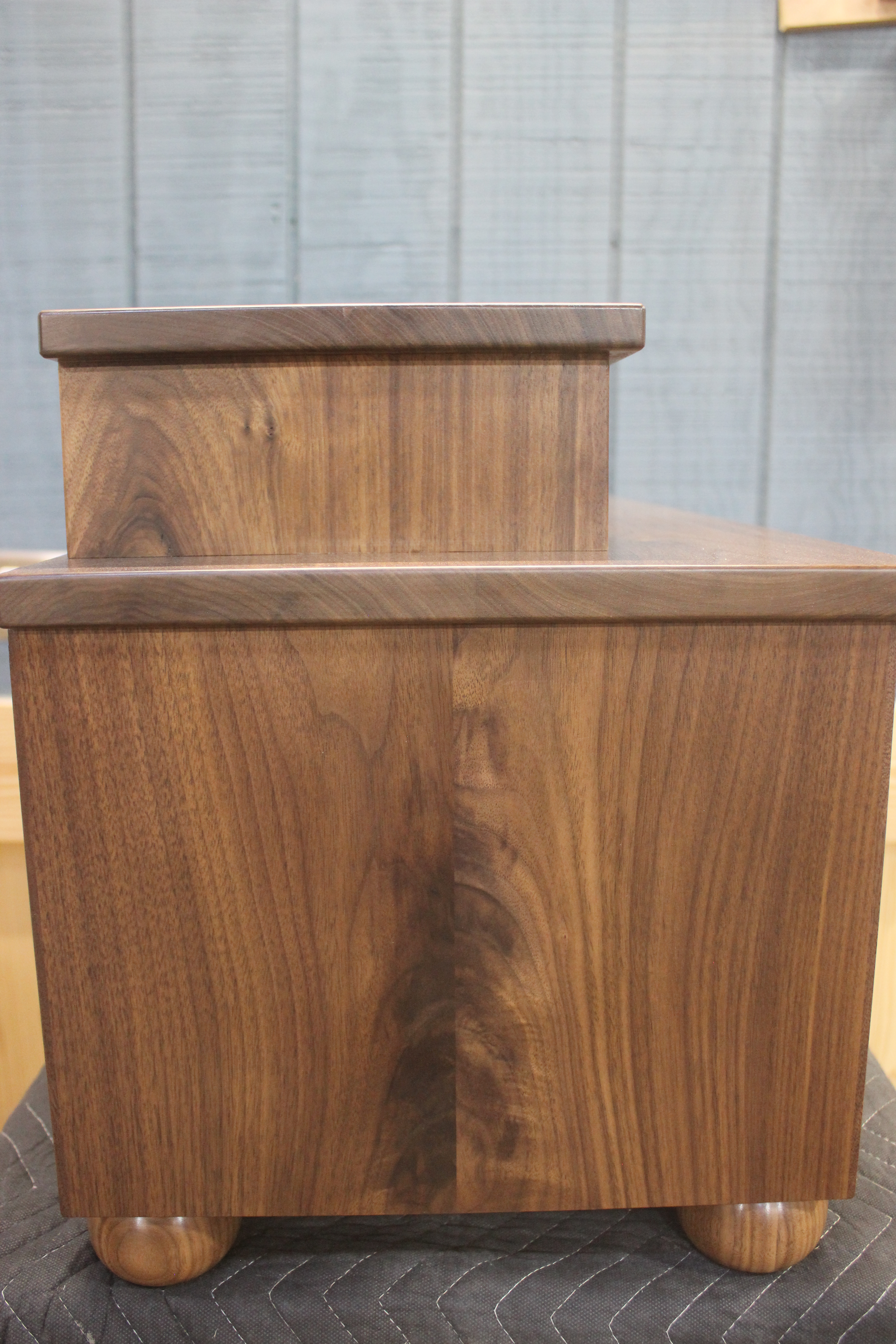 Side view of a wooden storage box with a smooth finish, featuring a raised top section and round wooden feet.