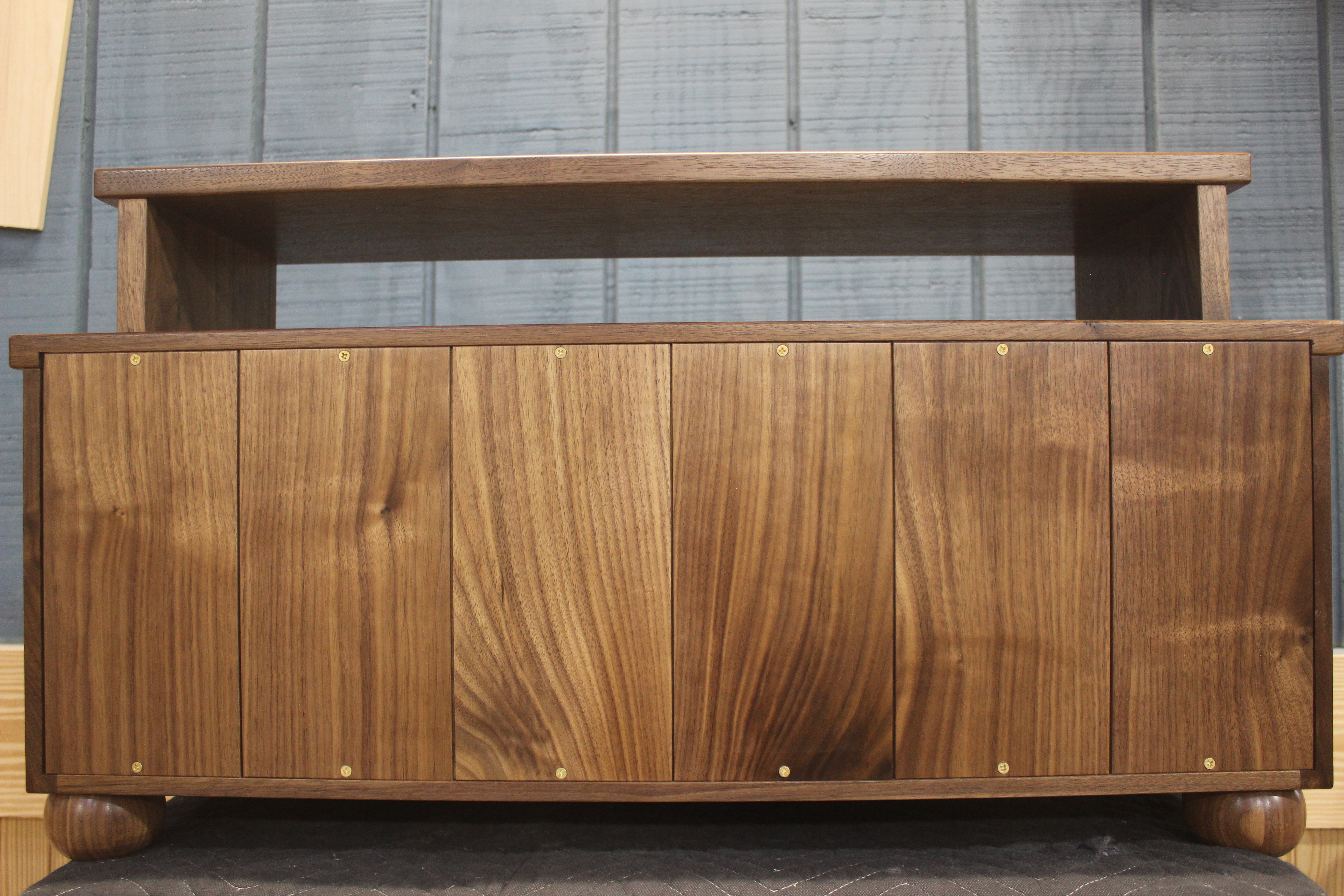 A modern wooden cabinet with a smooth, dark walnut finish. The cabinet features a top shelf and four front doors with brass handles, showcasing a sleek design.