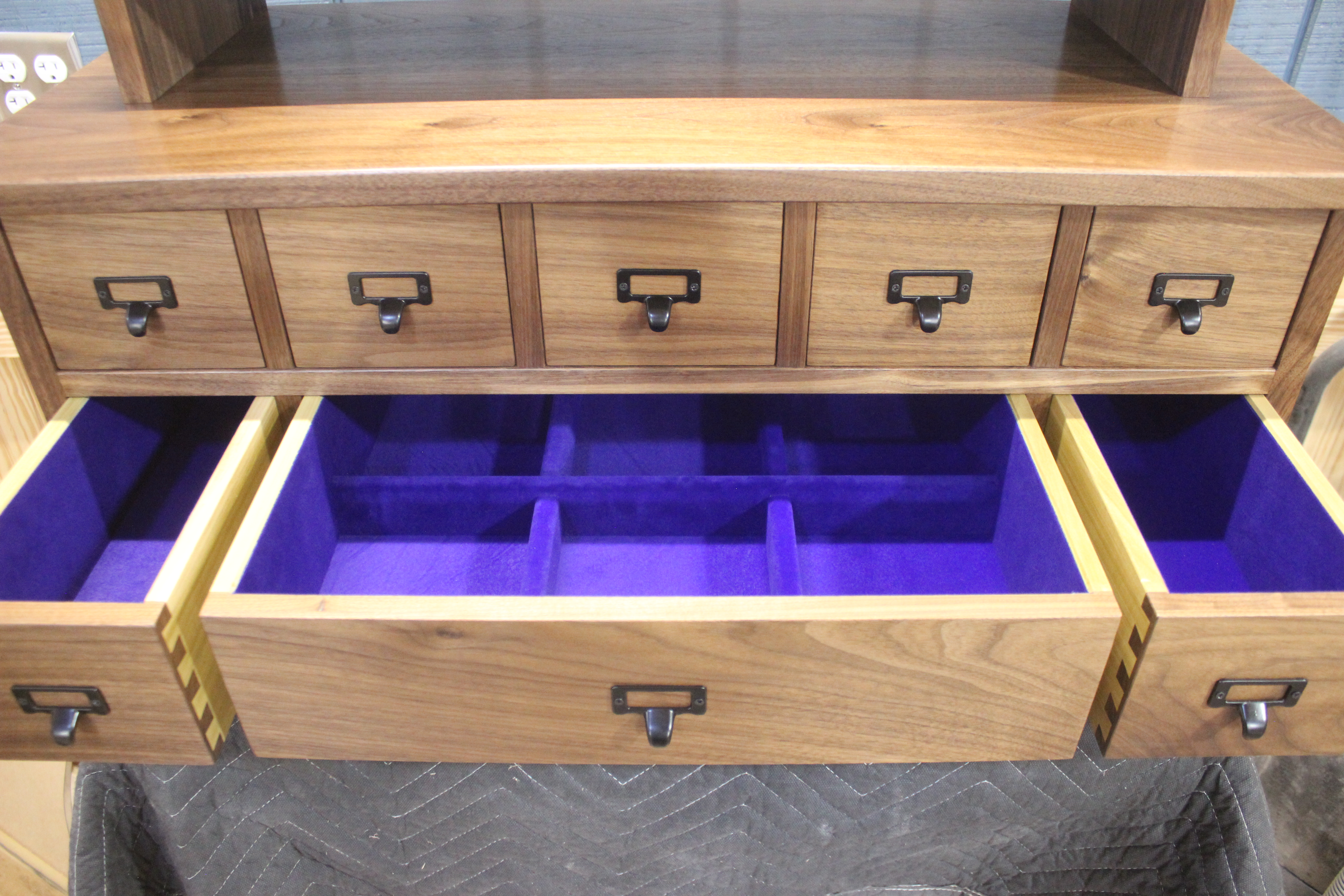 Close-up of a wooden cabinet featuring several small drawers with black handles, with one large drawer pulled out, revealing purple velvet lining and compartments.