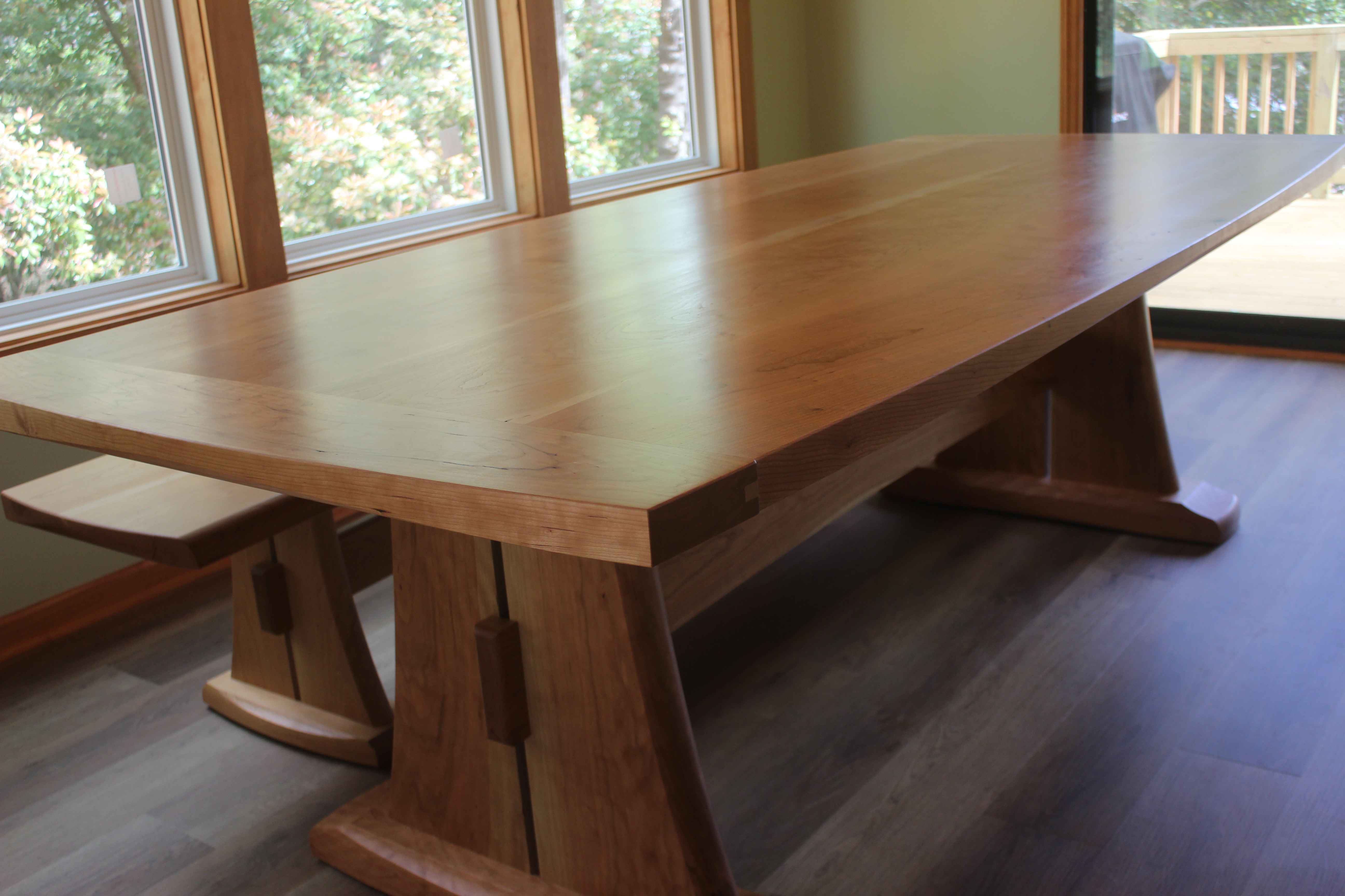 A large wooden dining table with a smooth surface and angled wooden legs, accompanied by a matching bench, situated near large windows that allow natural light to illuminate the space.