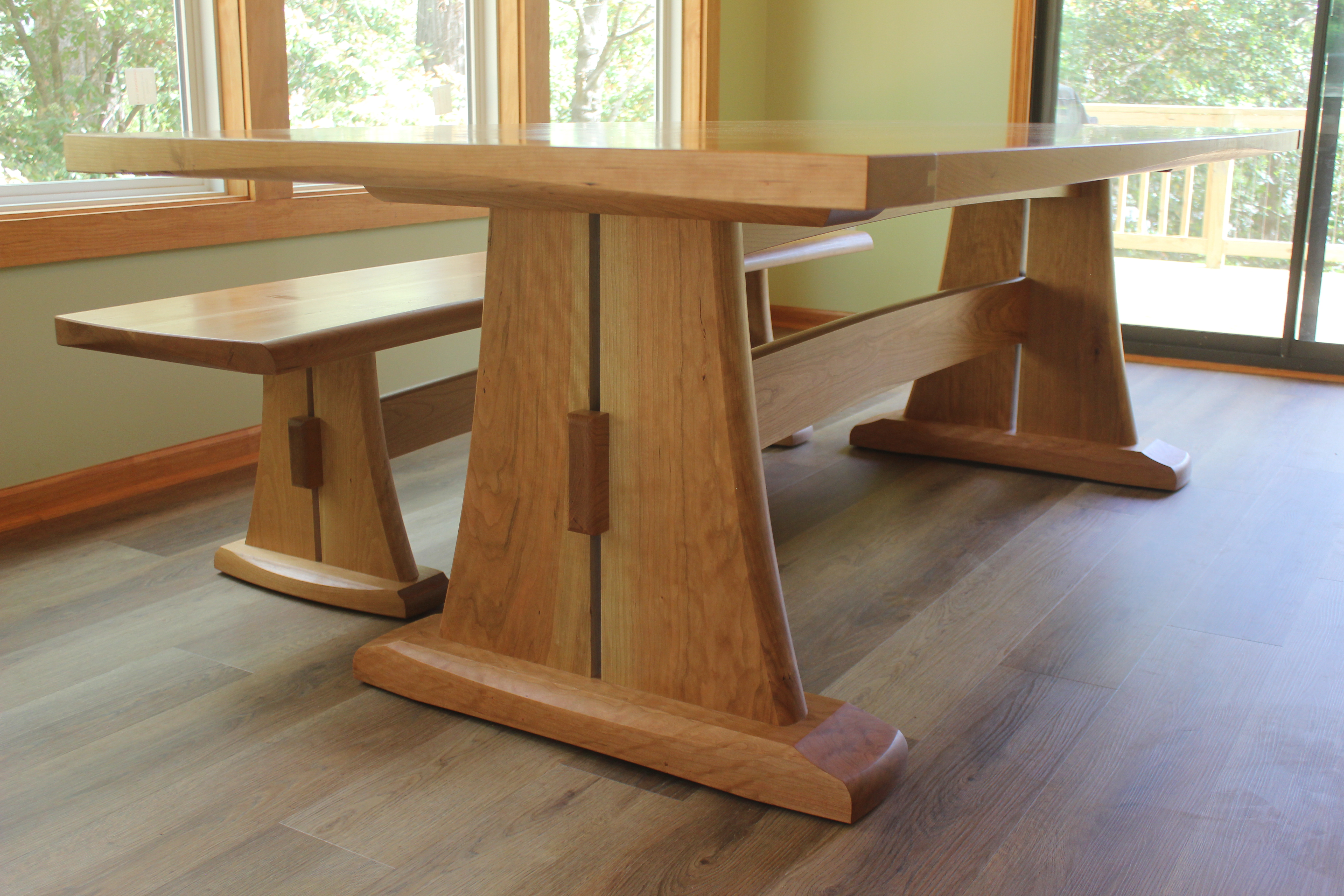 A modern wooden dining table with a matching bench, featuring a sturdy design with angled legs and a smooth surface, positioned near large windows.