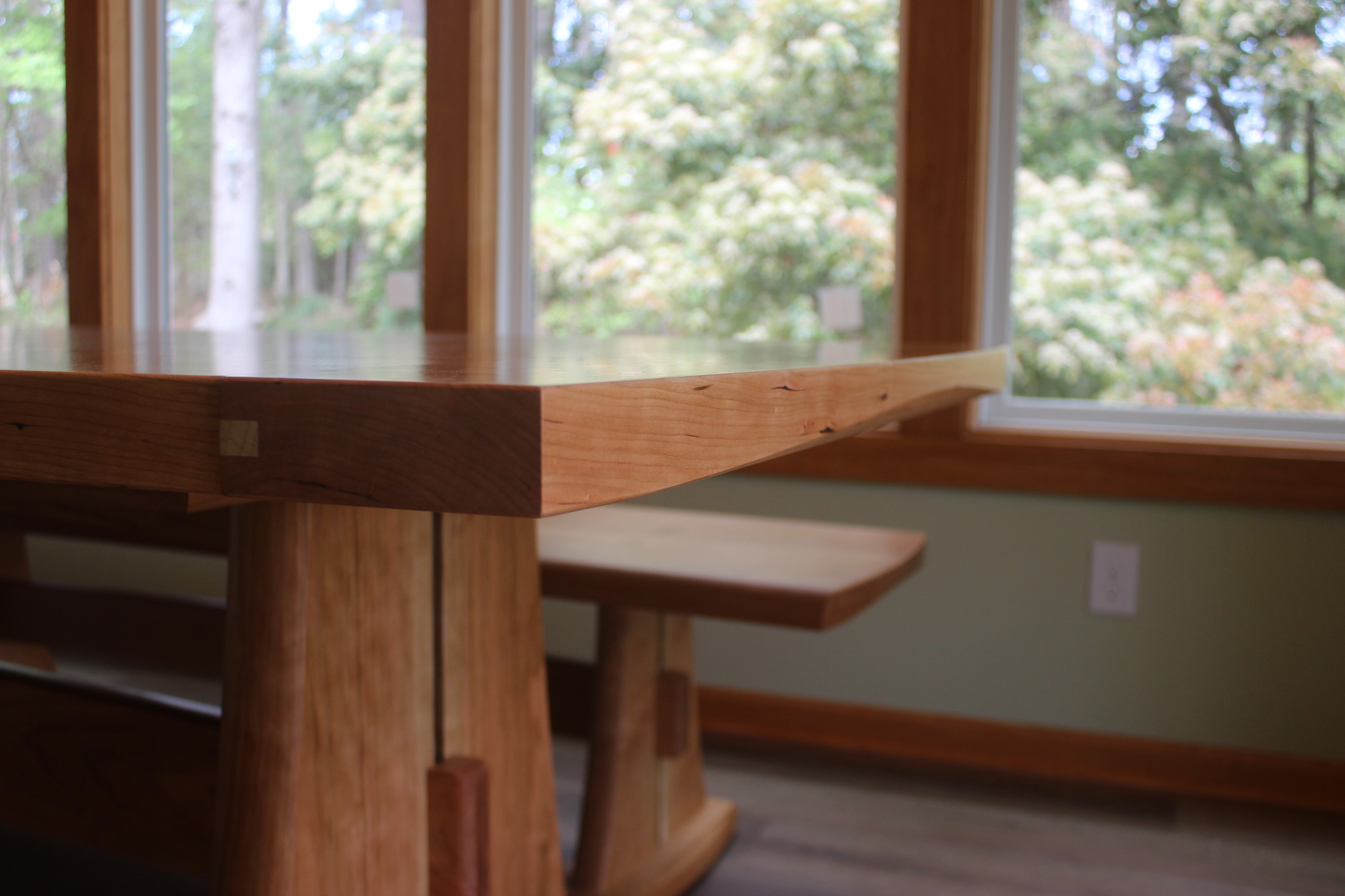 Close-up view of a wooden table with a smooth finish, showcasing its grain and craftsmanship, next to a bench and large windows with a green outdoor backdrop.