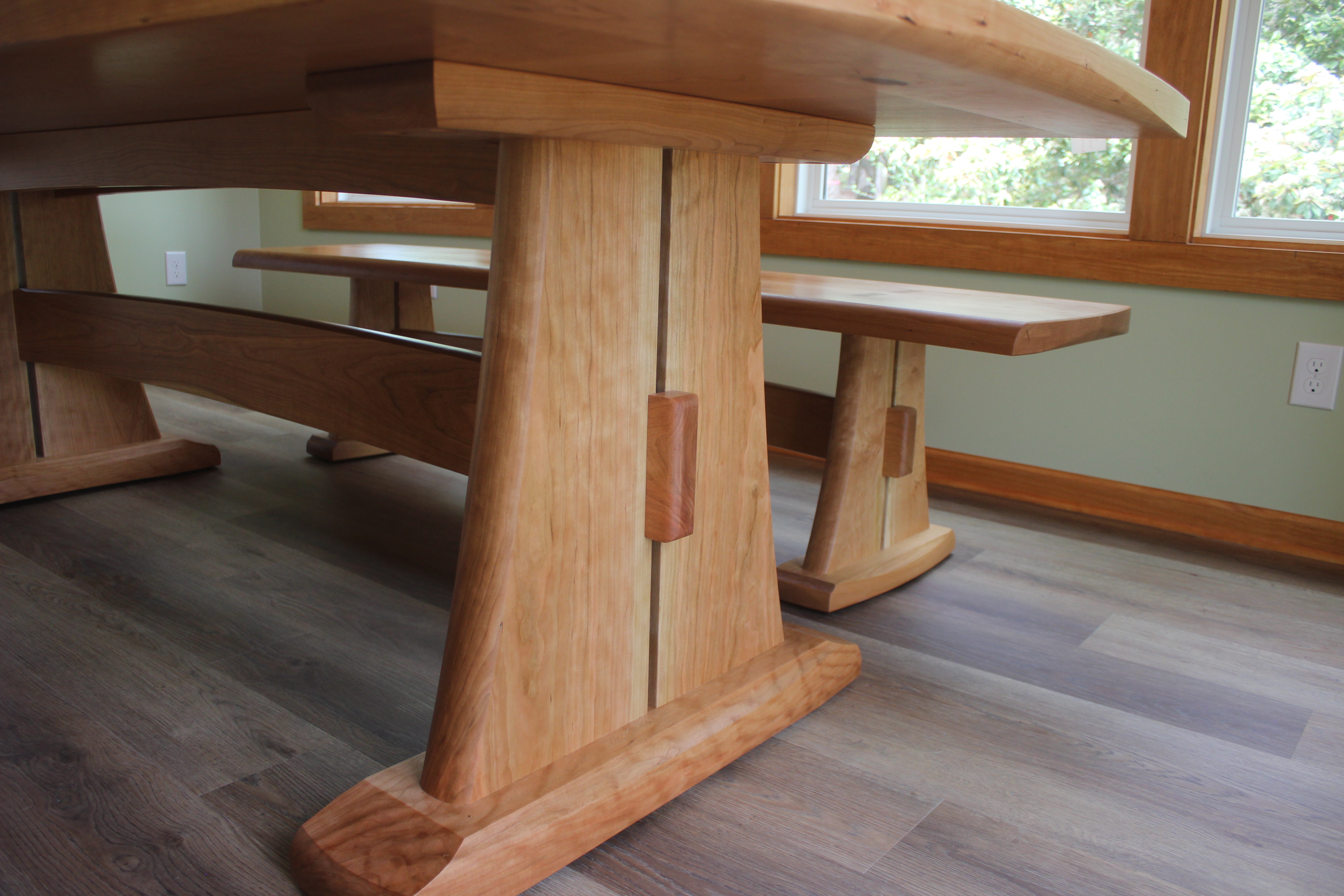 A close-up view of a wooden dining table and bench featuring a sleek design with sturdy legs and a polished surface, situated in a well-lit room with large windows.