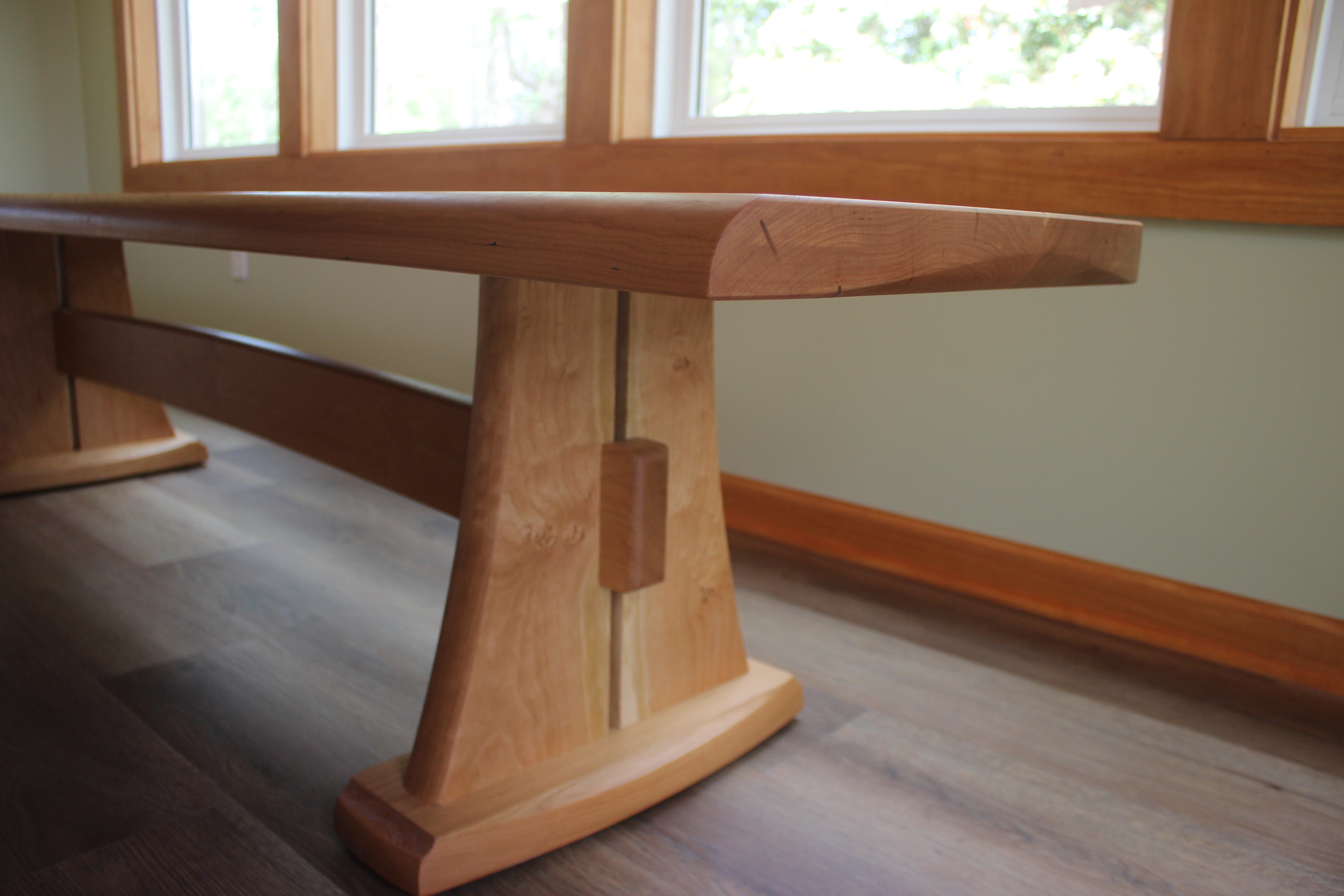 A wooden table with a smooth, curved surface and sturdy, uniquely shaped legs, set in a well-lit room with large windows.
