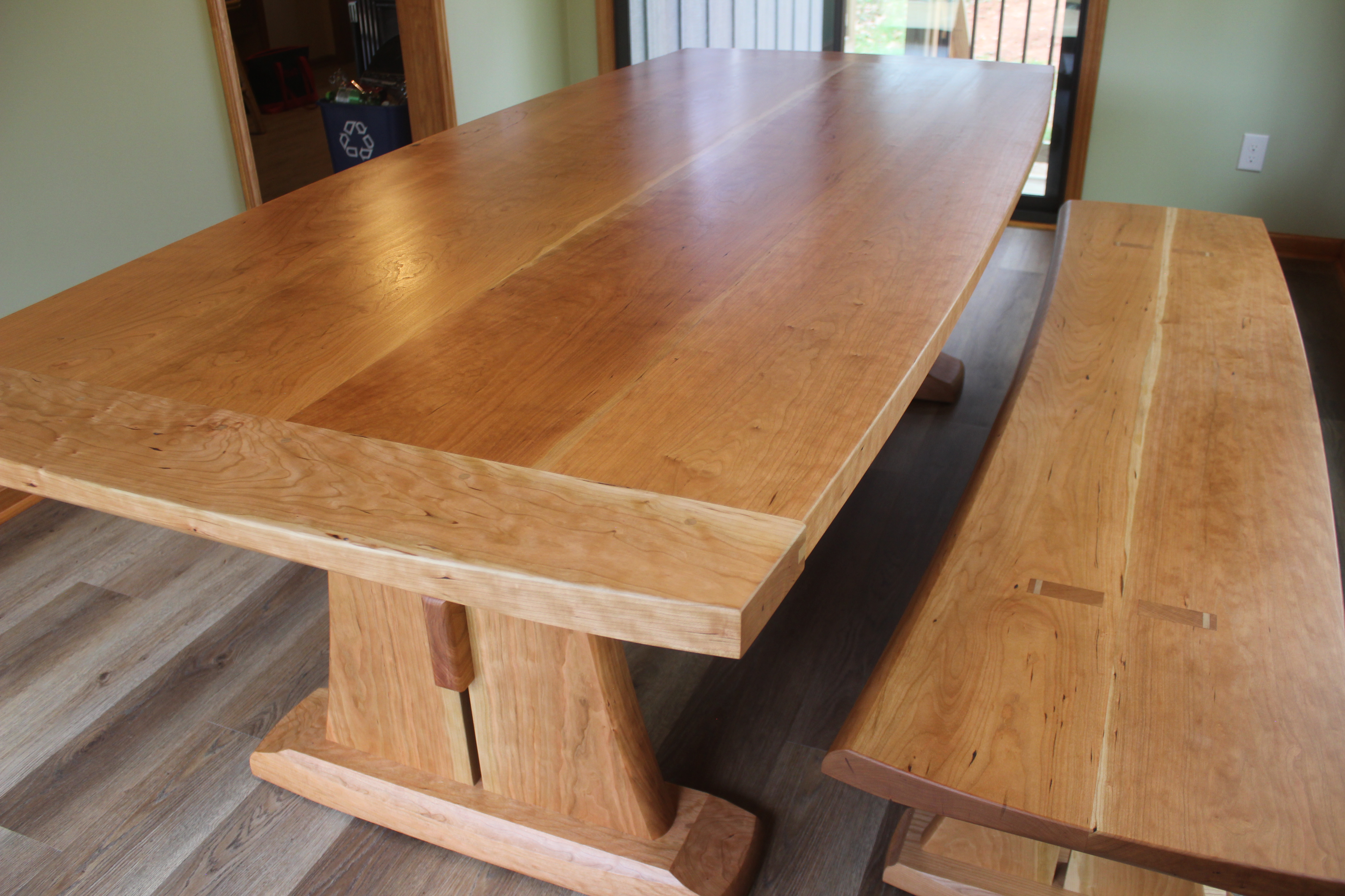 A modern wooden dining table with a smooth, light brown surface and matching bench, set in a bright indoor space.