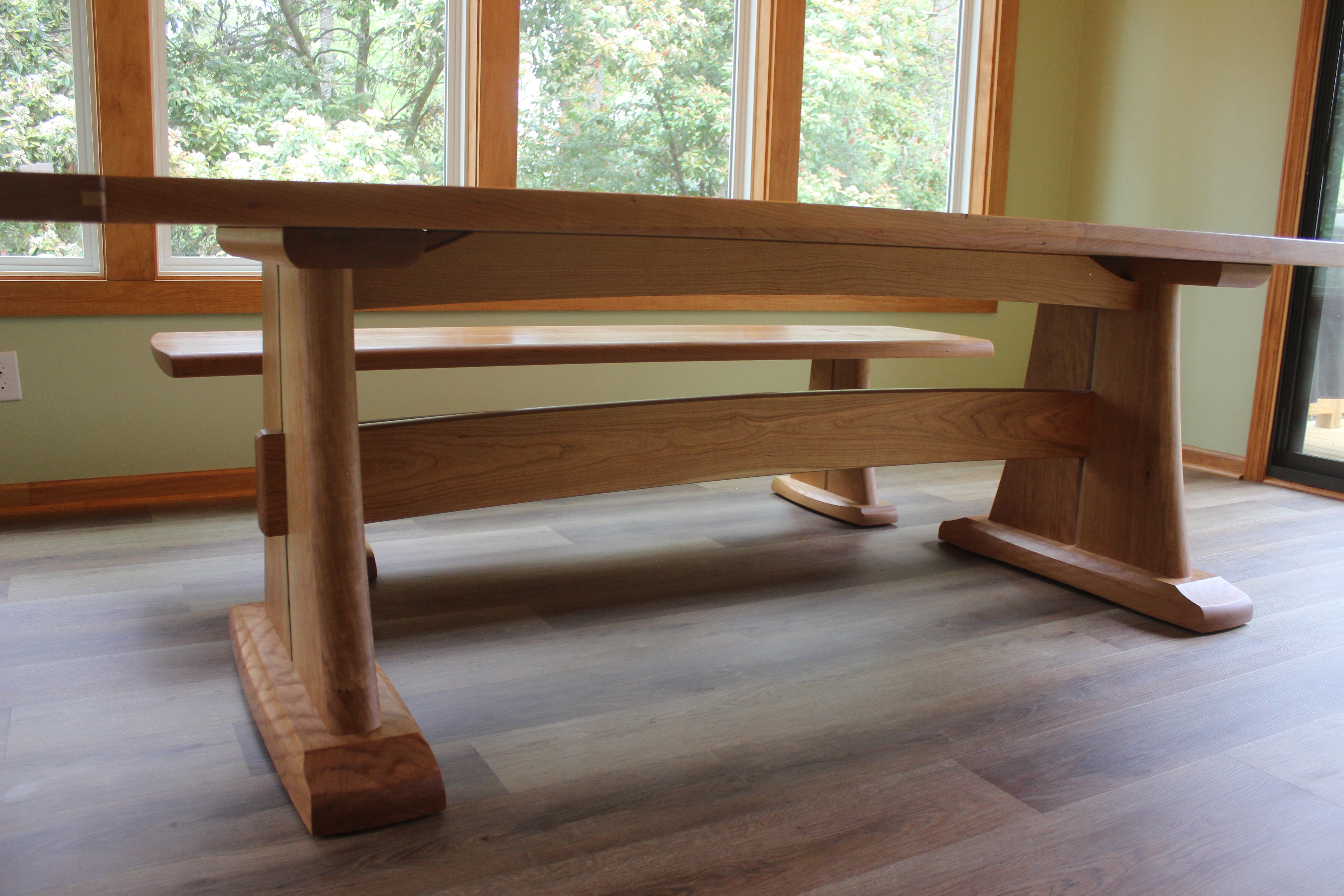 A handcrafted wooden dining table with a solid plank top and sturdy, uniquely designed legs, placed in a well-lit room with large windows and a green wall.