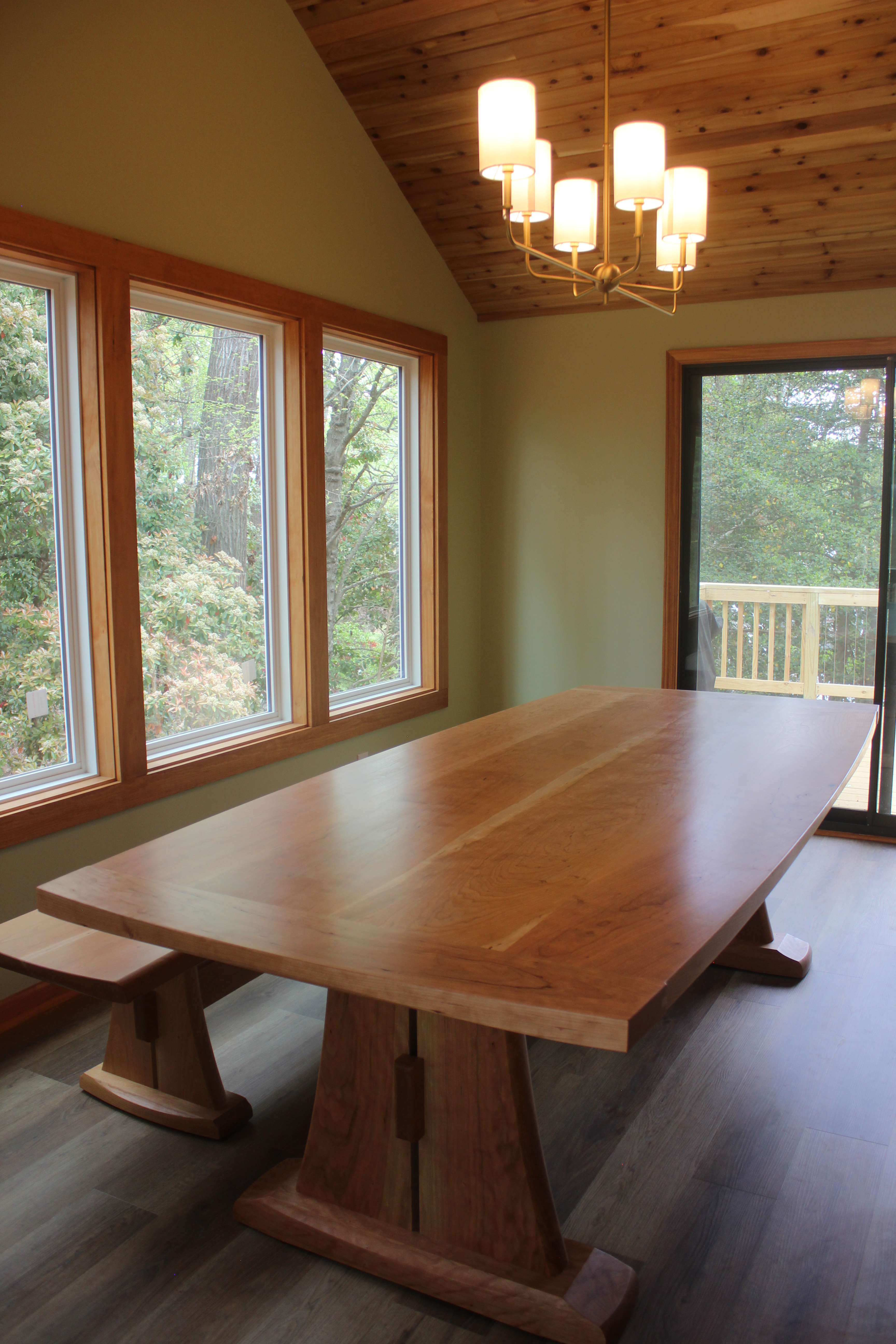 A spacious dining area featuring a large wooden table with sturdy legs, accompanied by a matching bench. The room has large windows providing natural light and a view of greenery outside, with a modern chandelier hanging from a wooden ceiling.