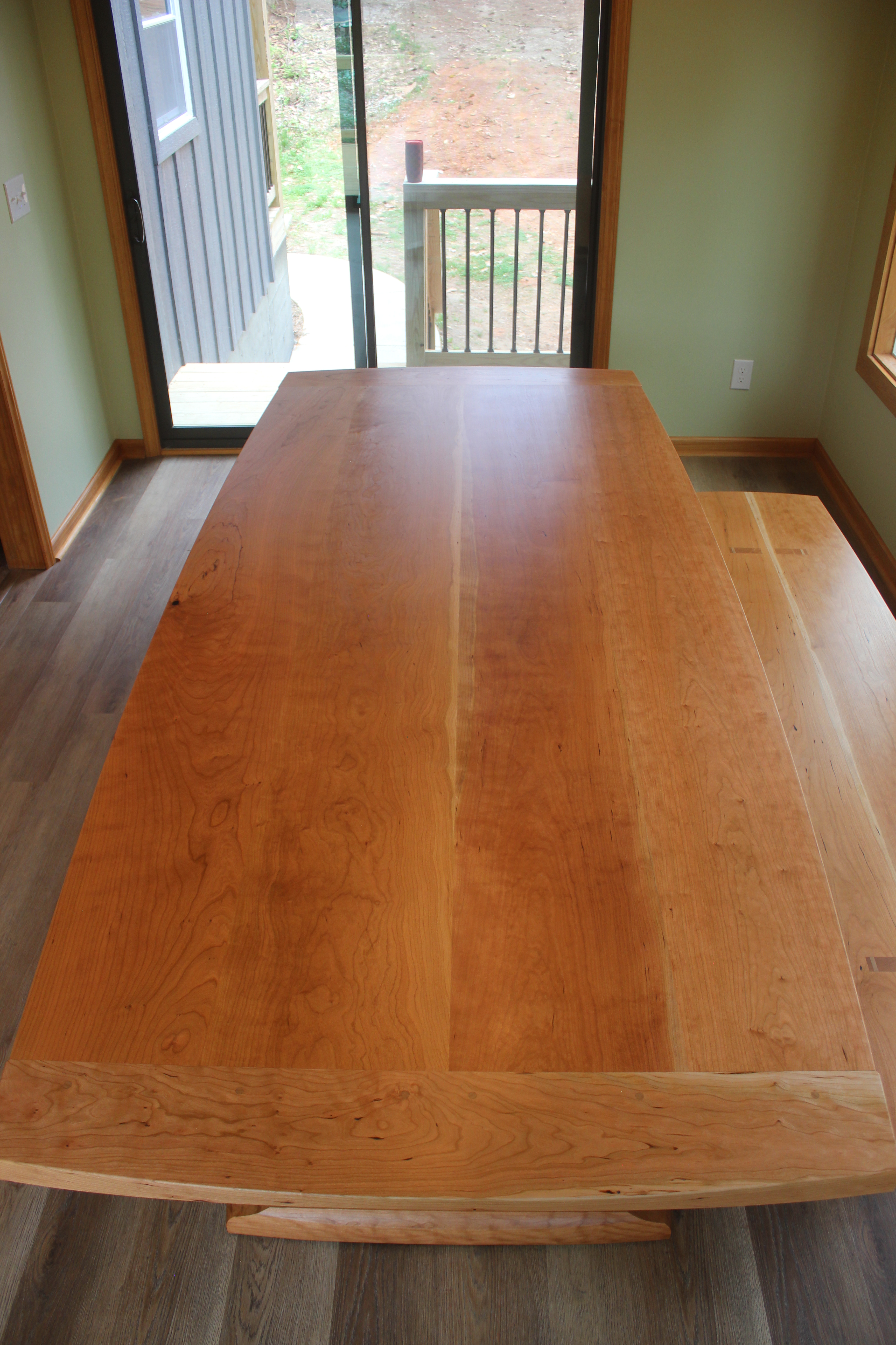A wooden dining table made of light-colored wood, viewed from above with a smooth surface, placed near a sliding glass door leading outside.