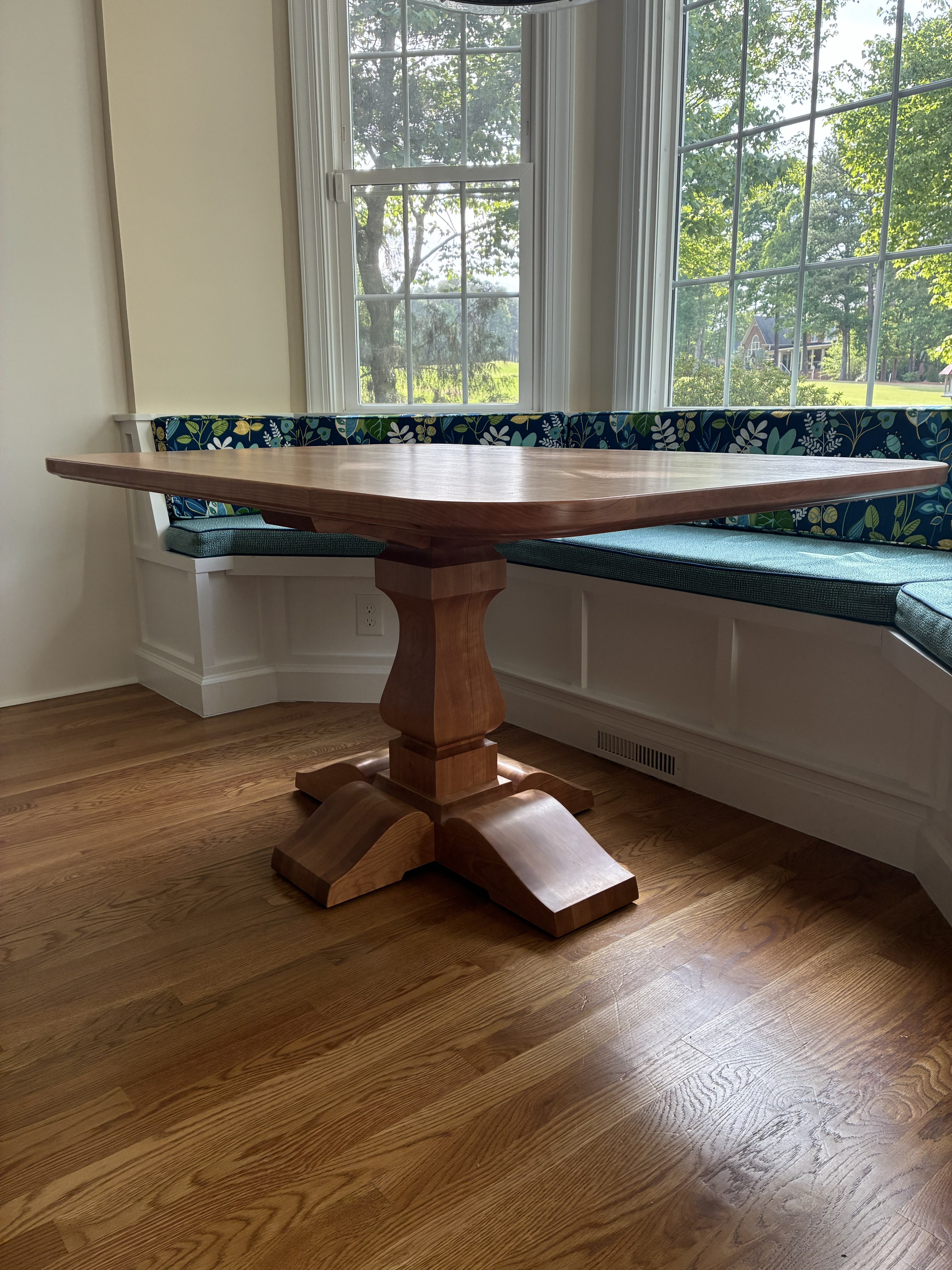 A wooden dining table with a unique pedestal base, surrounded by large windows that let in natural light and a cozy seating area with blue floral cushions.