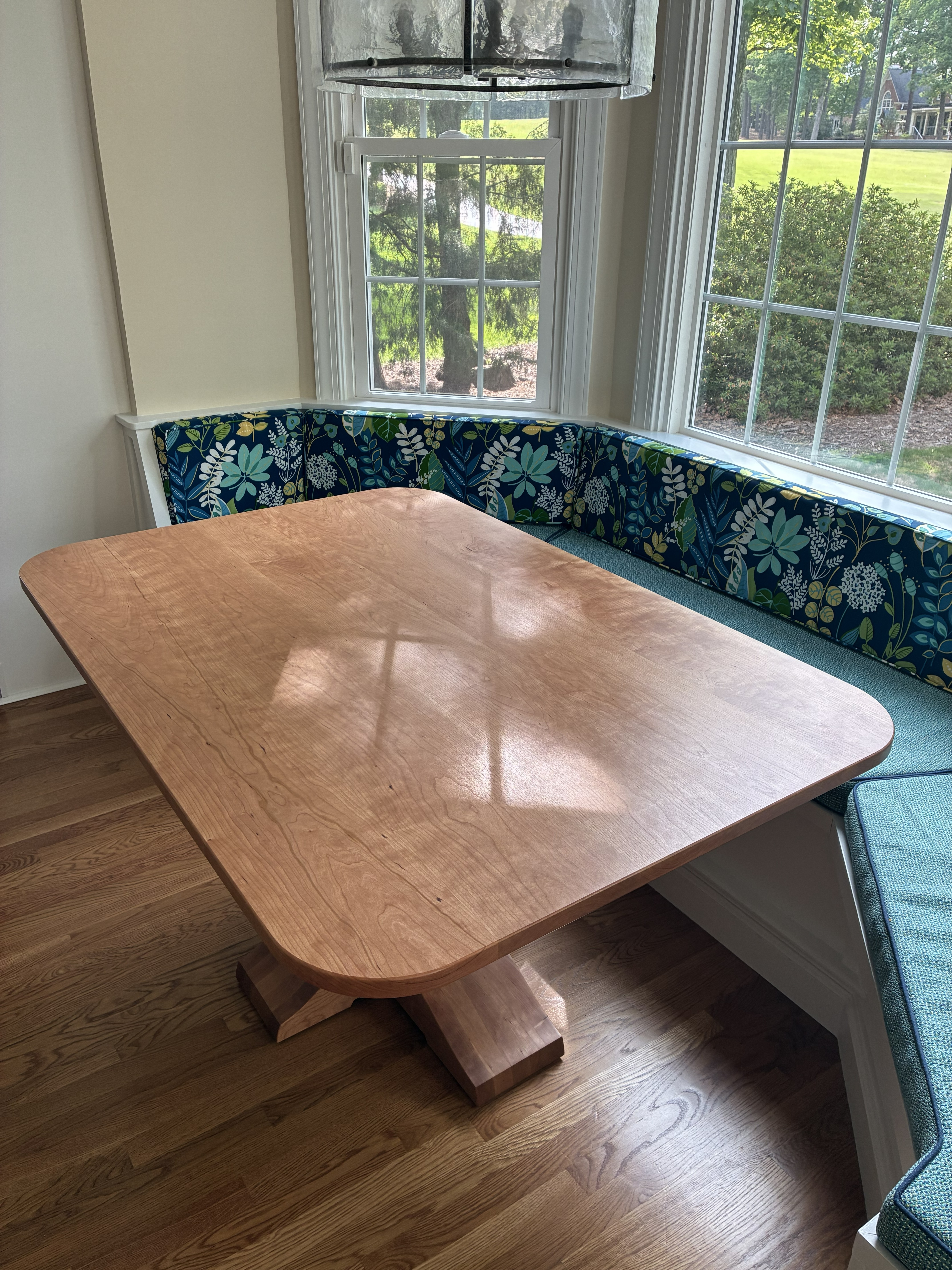 A wooden dining table with a smooth surface and unique base, positioned in a bright sunny area beside large windows. There is a floral-patterned cushioned bench along the walls.