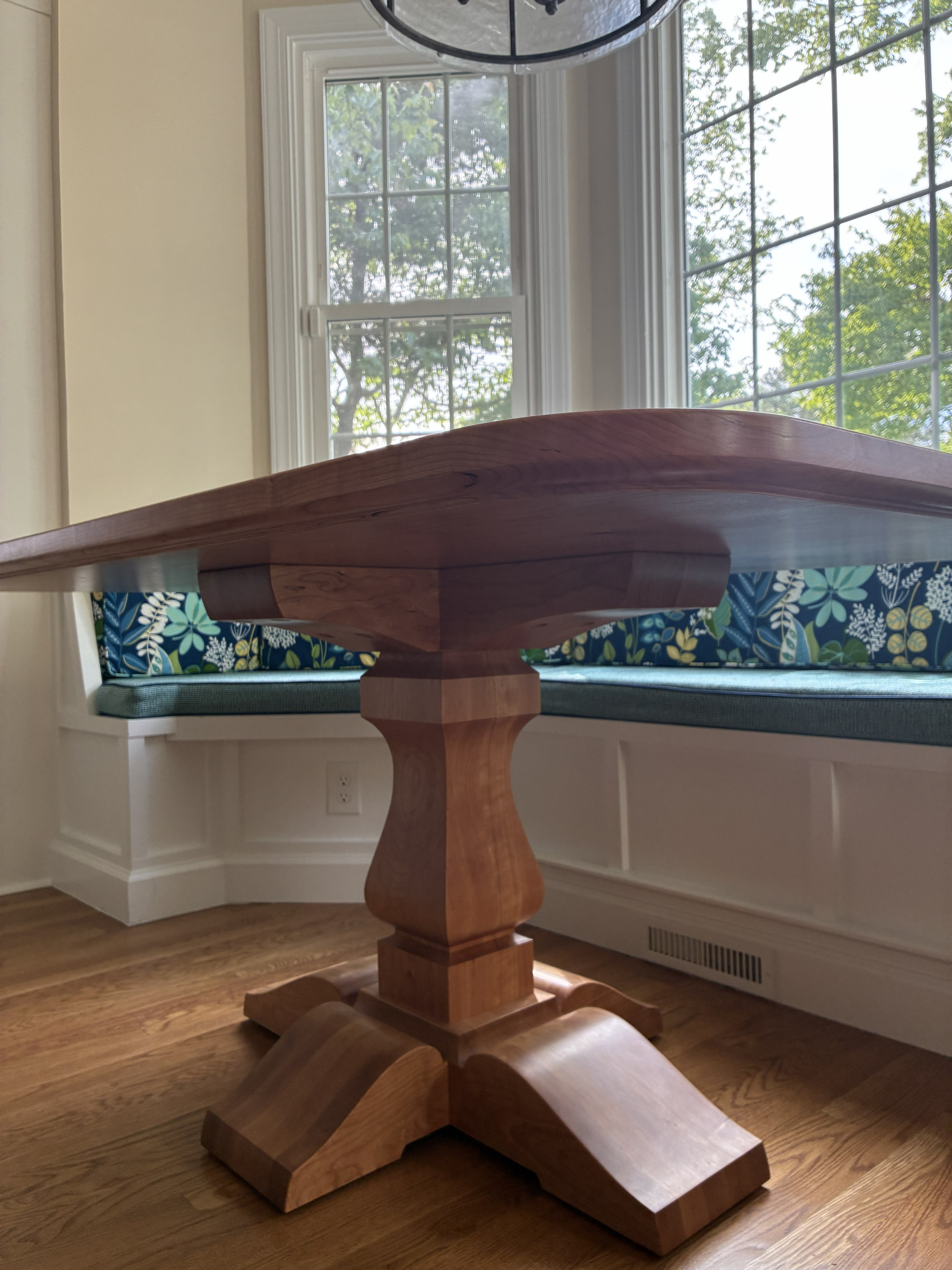 A wooden dining table with a polished surface and a decorative pedestal base, situated in a well-lit room with large windows and a cozy window seat adorned with floral cushions.