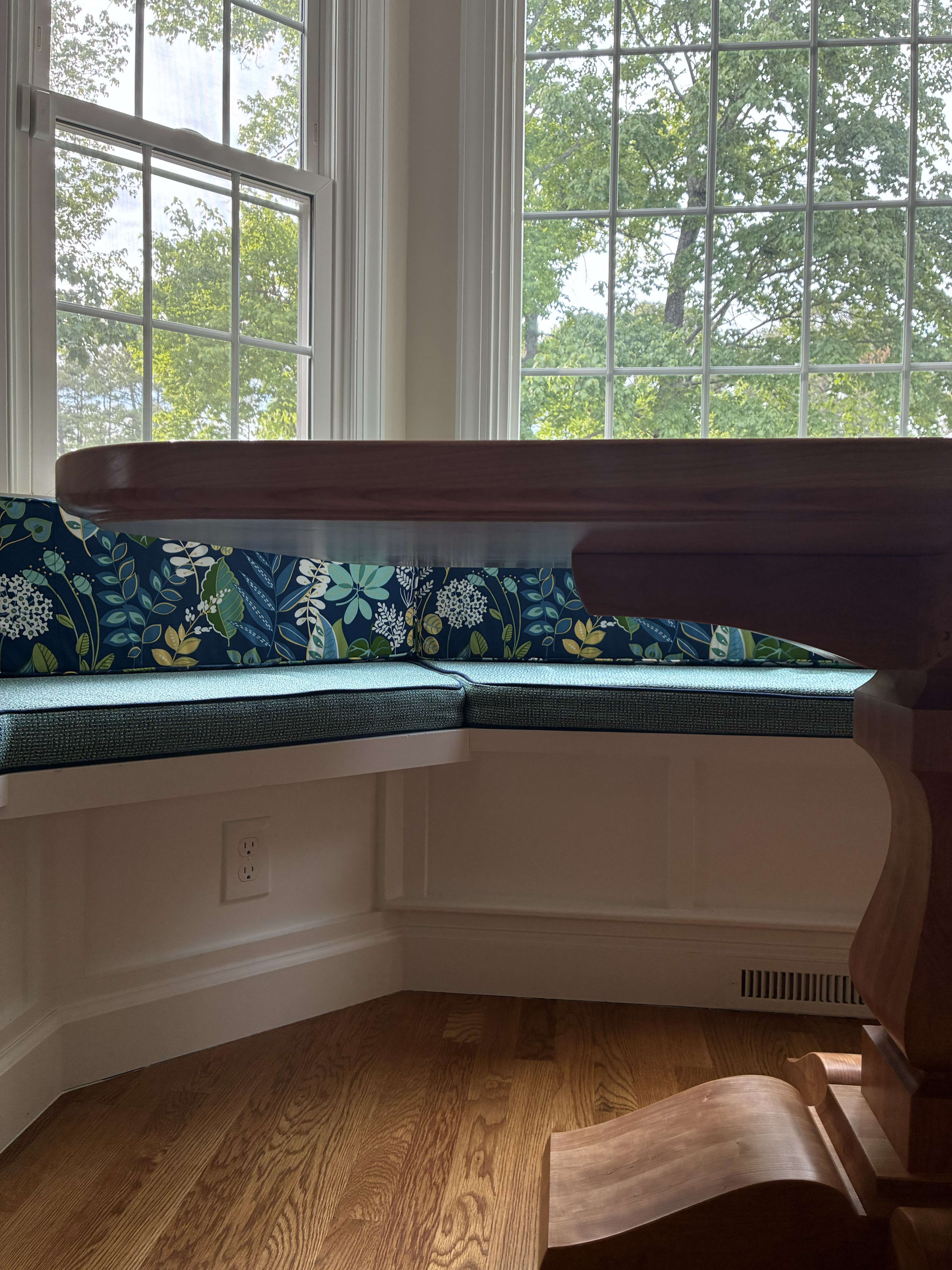 A cozy dining nook featuring a wooden table and floral-patterned cushions by the windows, surrounded by greenery.