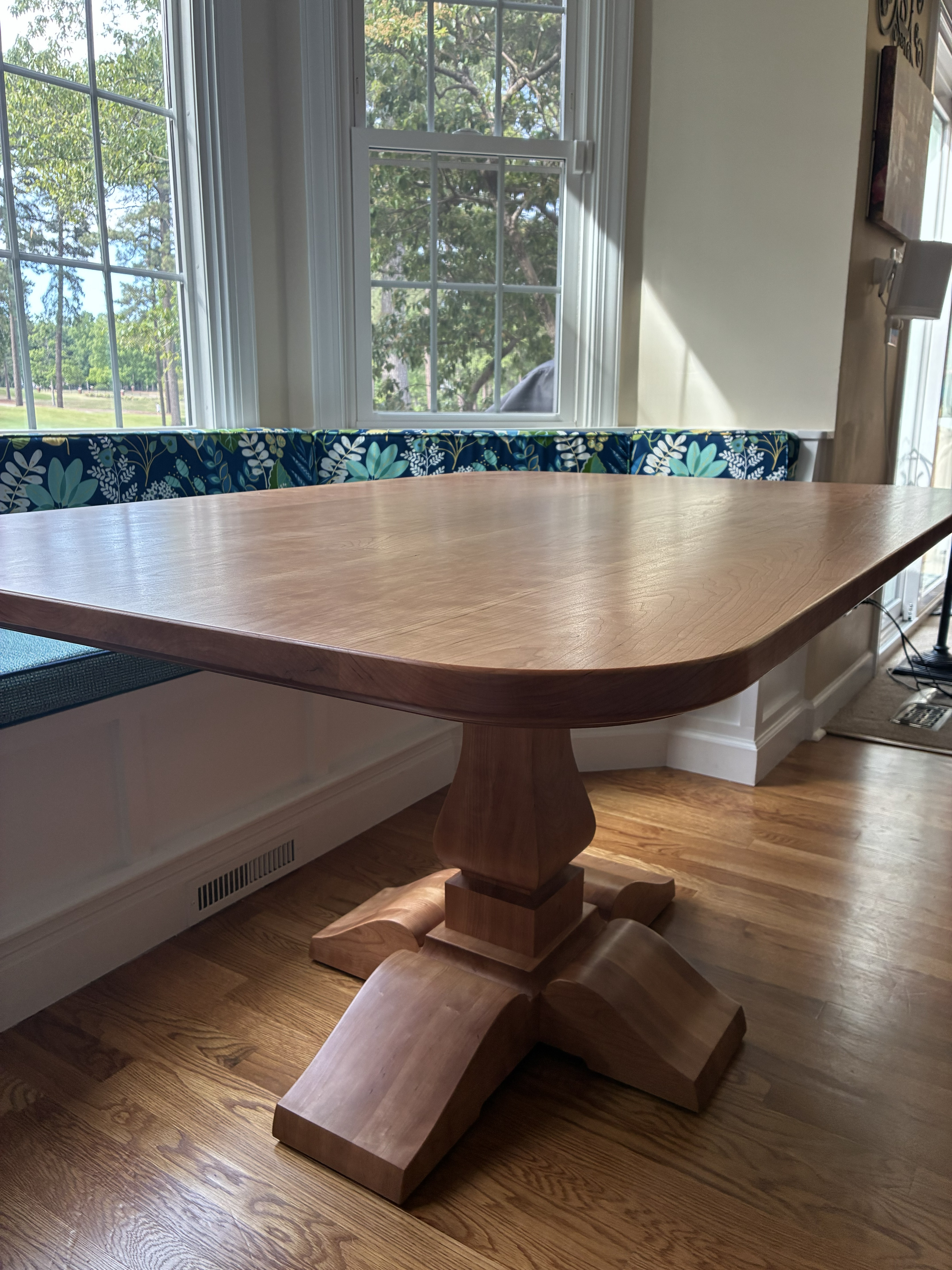 A wooden dining table with a rounded edge, supported by a decorative pedestal base, positioned near a window with a floral cushion on a nearby bench.