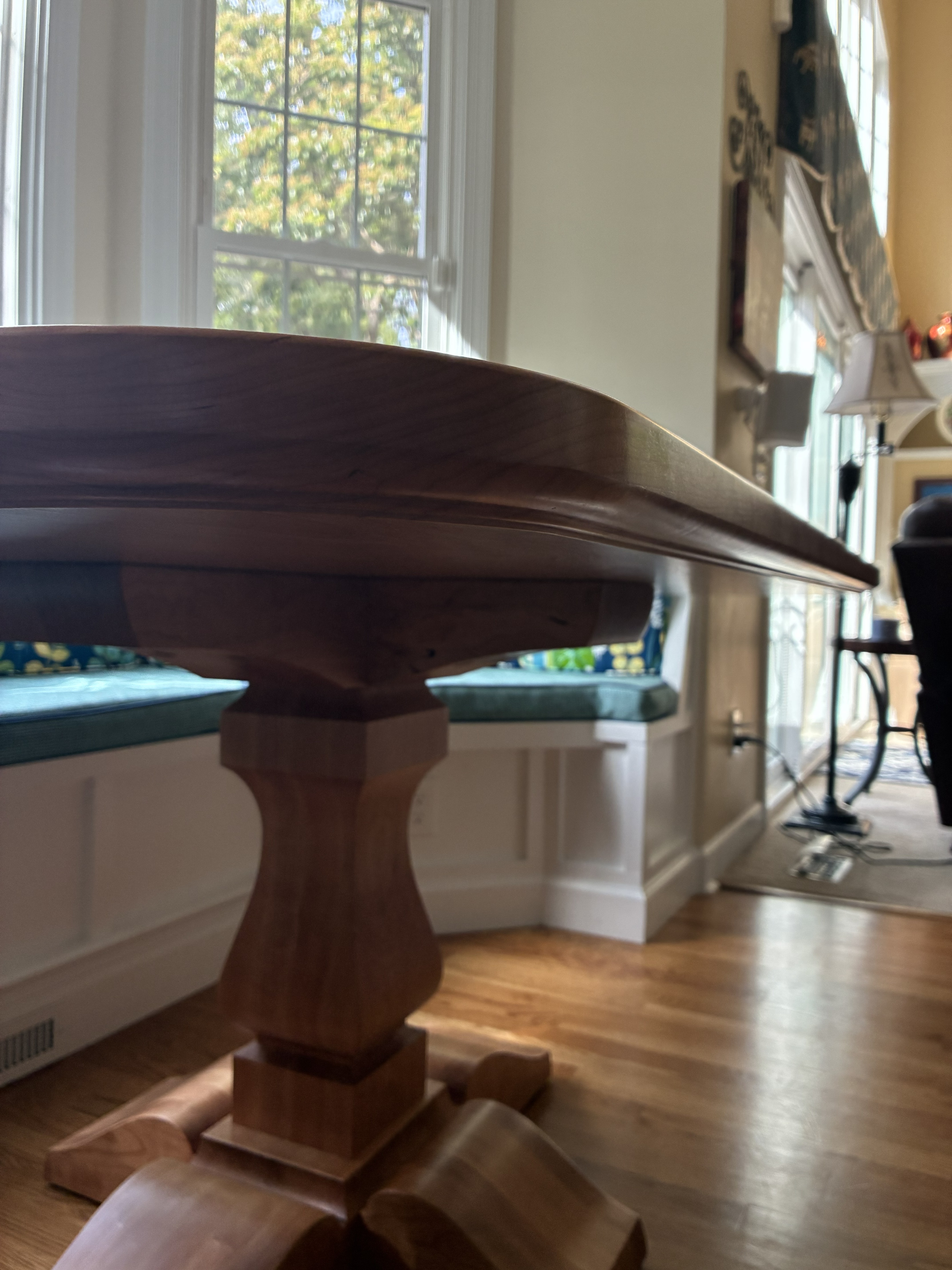 A close-up view of a wooden dining table with a detailed pedestal base, set in a bright room with large windows.